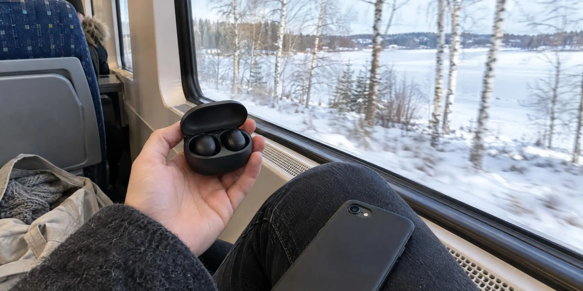 Laddfodral med trådlösa hörlurar i handen på ett tåg, med snötäckta björkar utanför fönstret