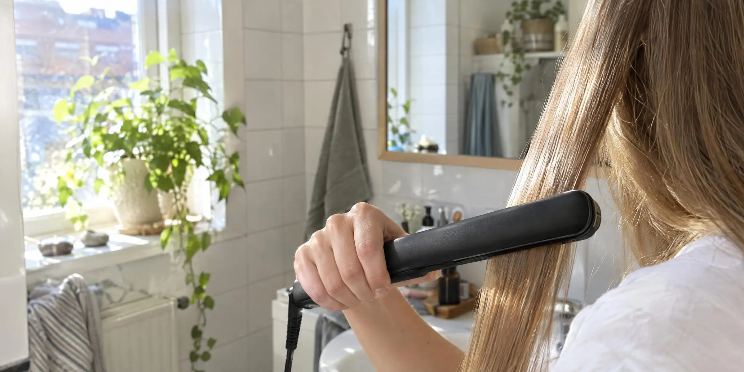 En hand plattar långt hår med en plattång framför spegeln i ett ljust badrum