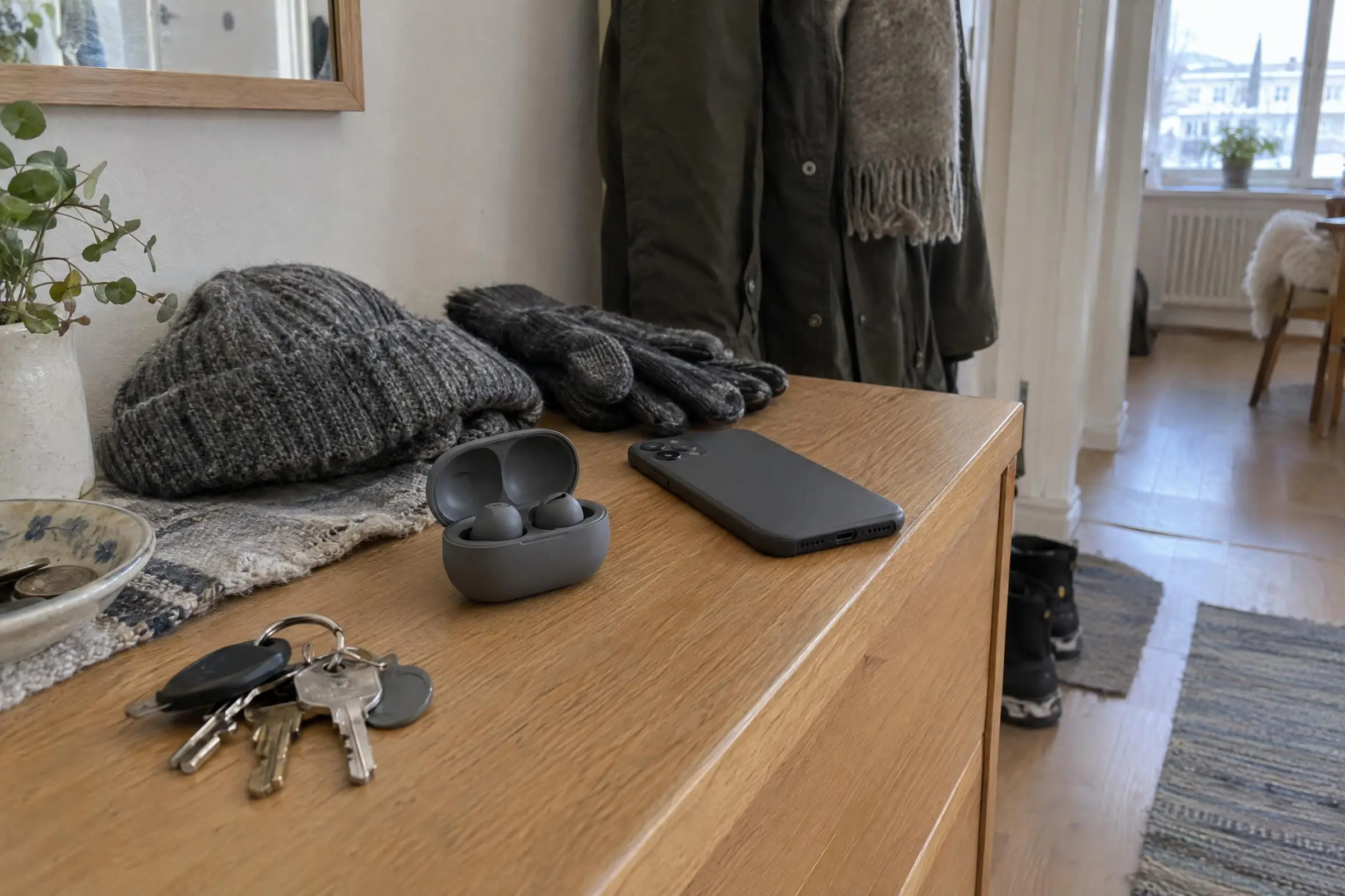 Ett öppet laddfodral med två trådlösa hörlurar på en hallbyrå, bredvid en mobil och en nyckelknippa.